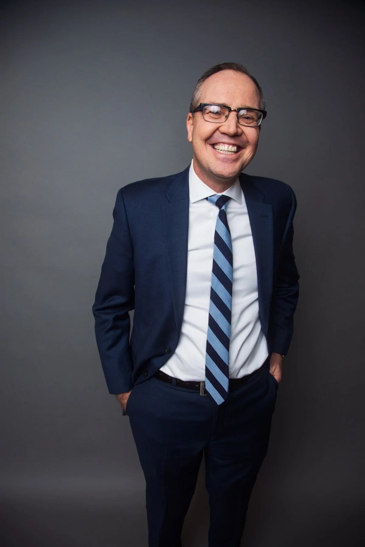 A smiling man in a dark blue suit, white shirt, and striped blue tie standing against a gray background.