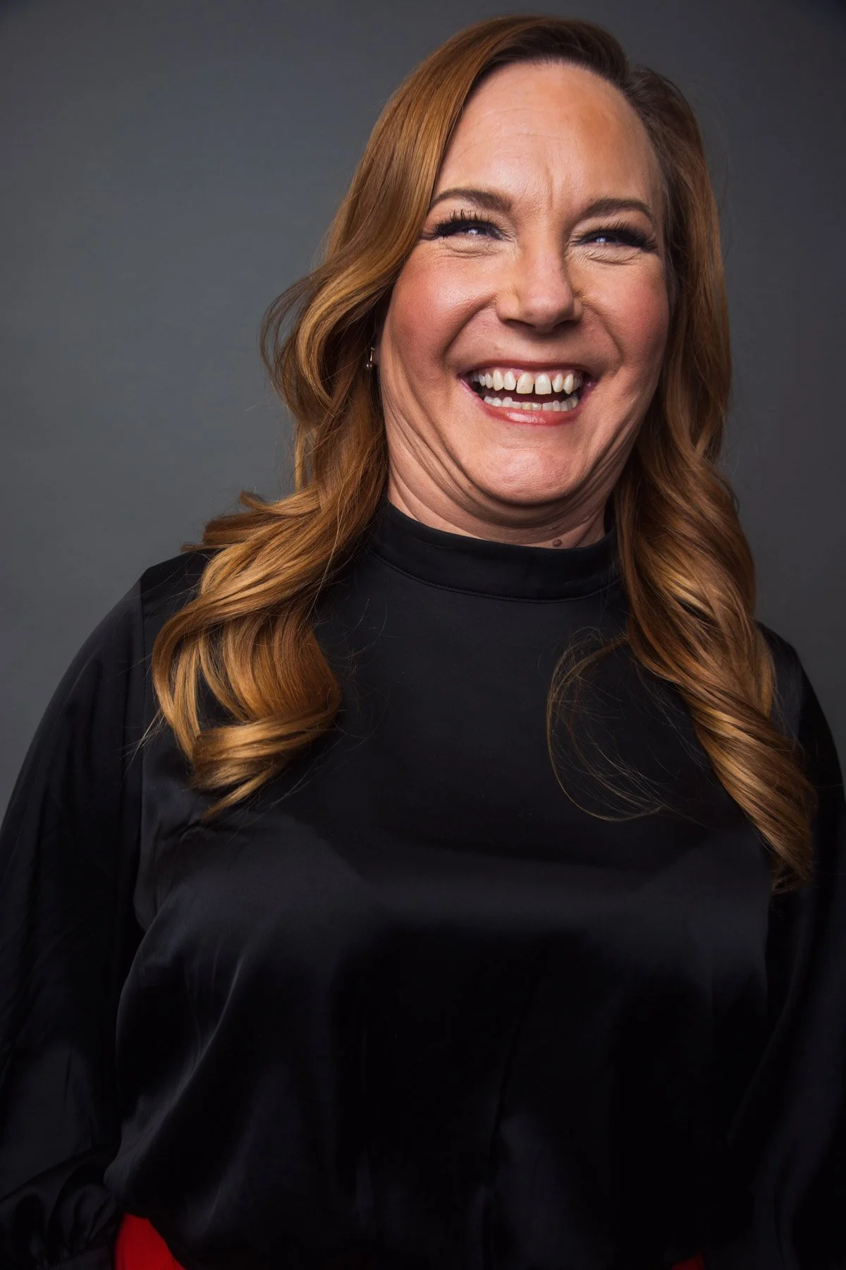A smiling woman with red hair wearing a black top with a high neckline, standing against a gray background.