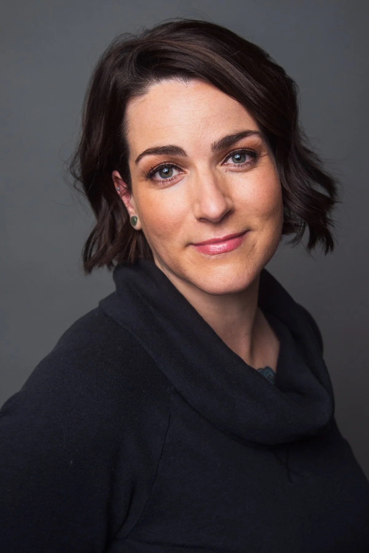 A woman with shoulder-length dark brown hair styled in loose waves, blue eyes, and light makeup, wearing a black top, smiling against a grey background.