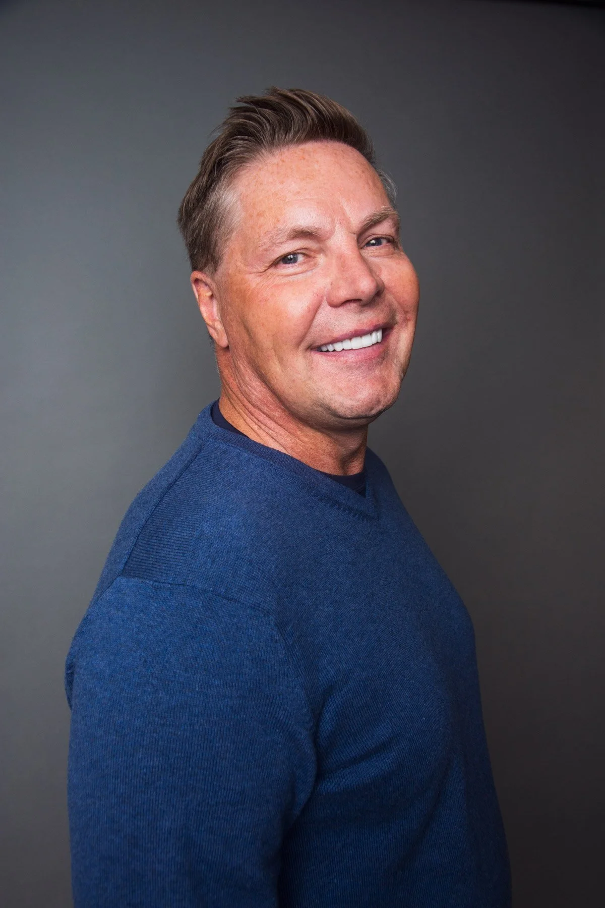 A smiling man with short brown hair wearing a blue sweater, standing against a gray background.
