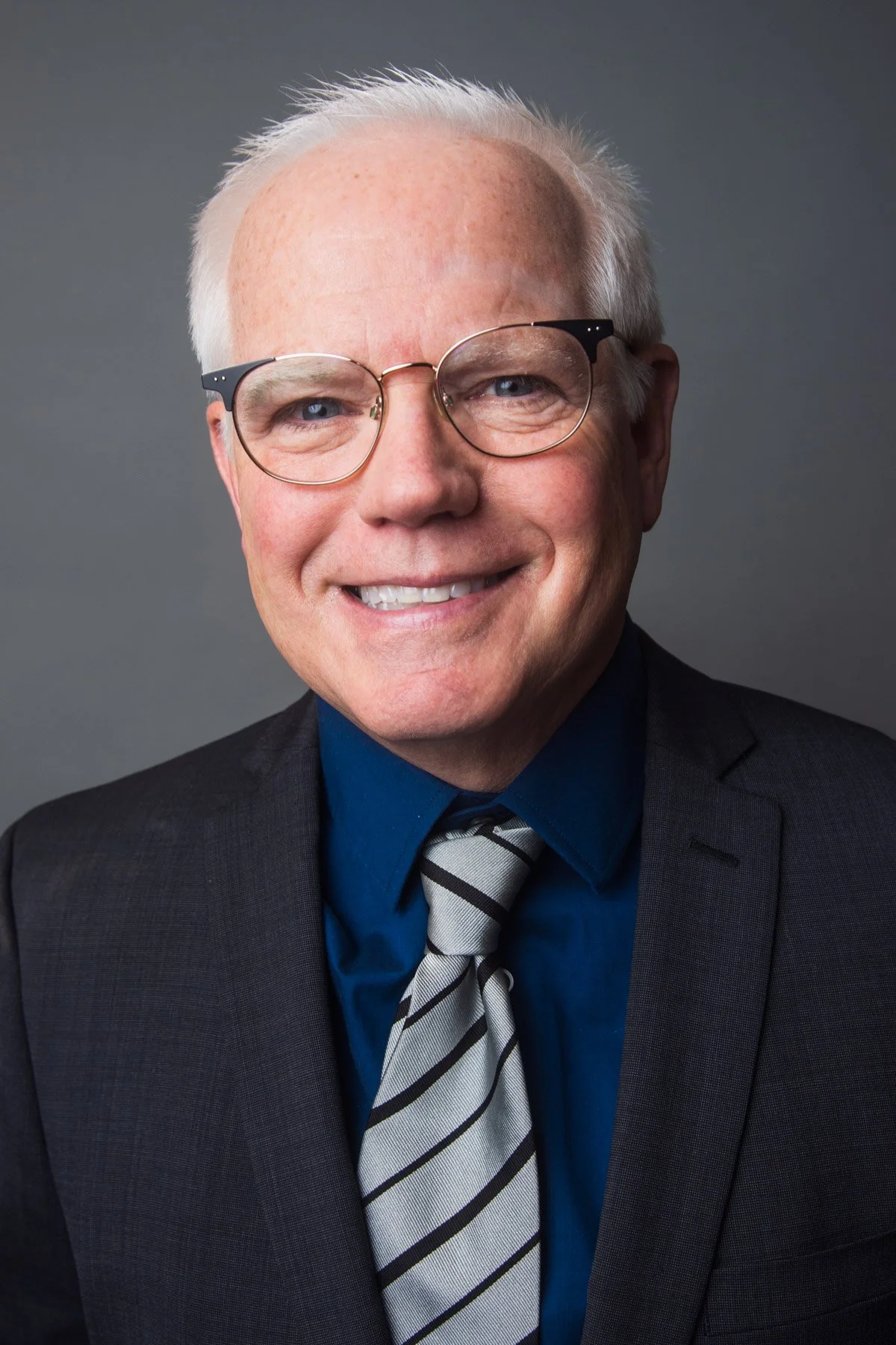 A smiling older man with white hair and glasses, wearing a black suit, blue shirt, and striped tie, against a gray background.