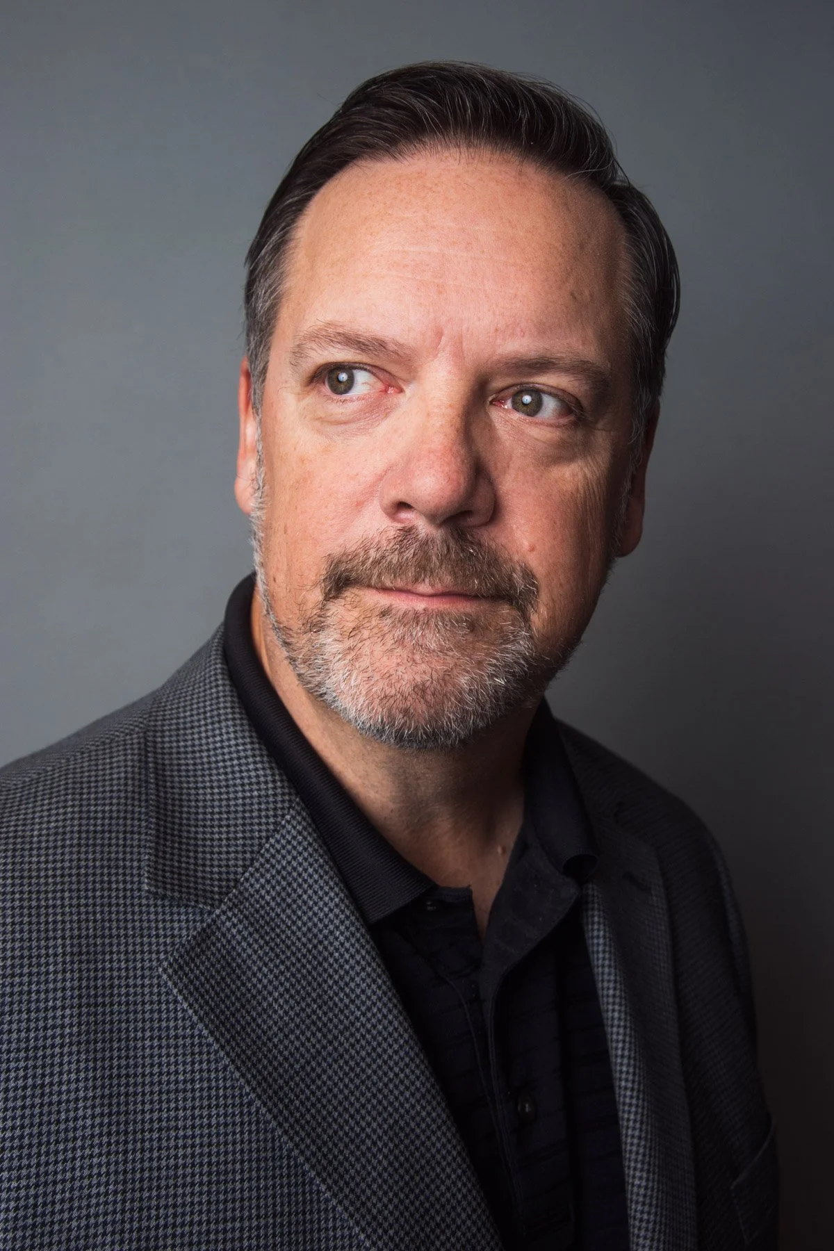 A middle-aged man with dark hair, a mustache and beard, wearing a patterned blazer and black shirt against a gray background.