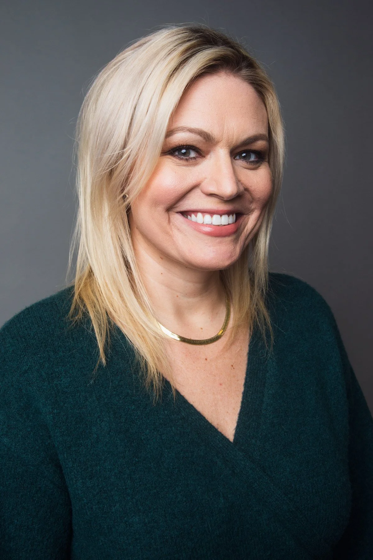 A smiling woman with blonde hair, blue eyes, and freckles, wearing a dark green sweater and a gold necklace, against a gray background.