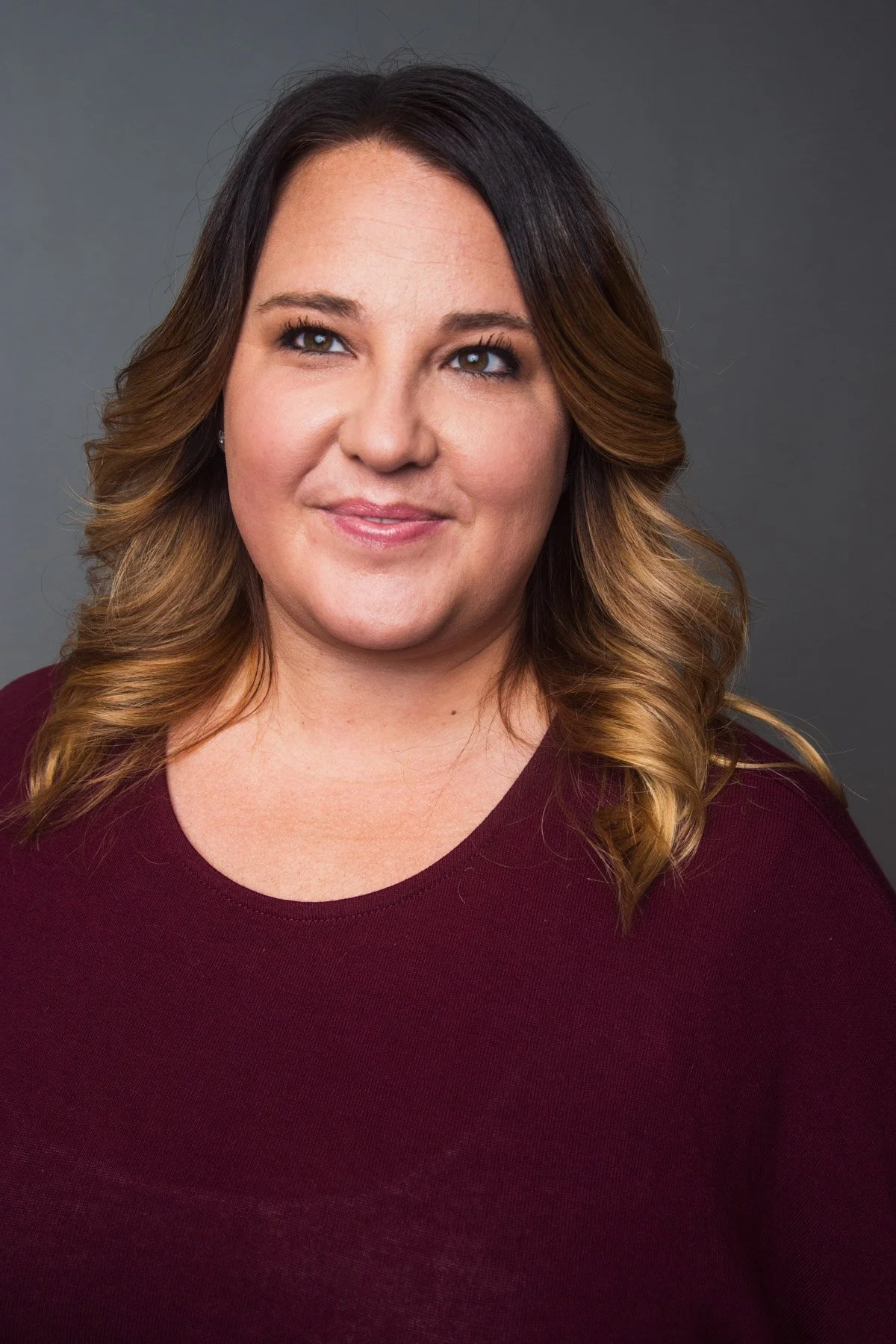 A woman with shoulder-length wavy hair and a slight smile wearing a maroon top, posed against a grey background.