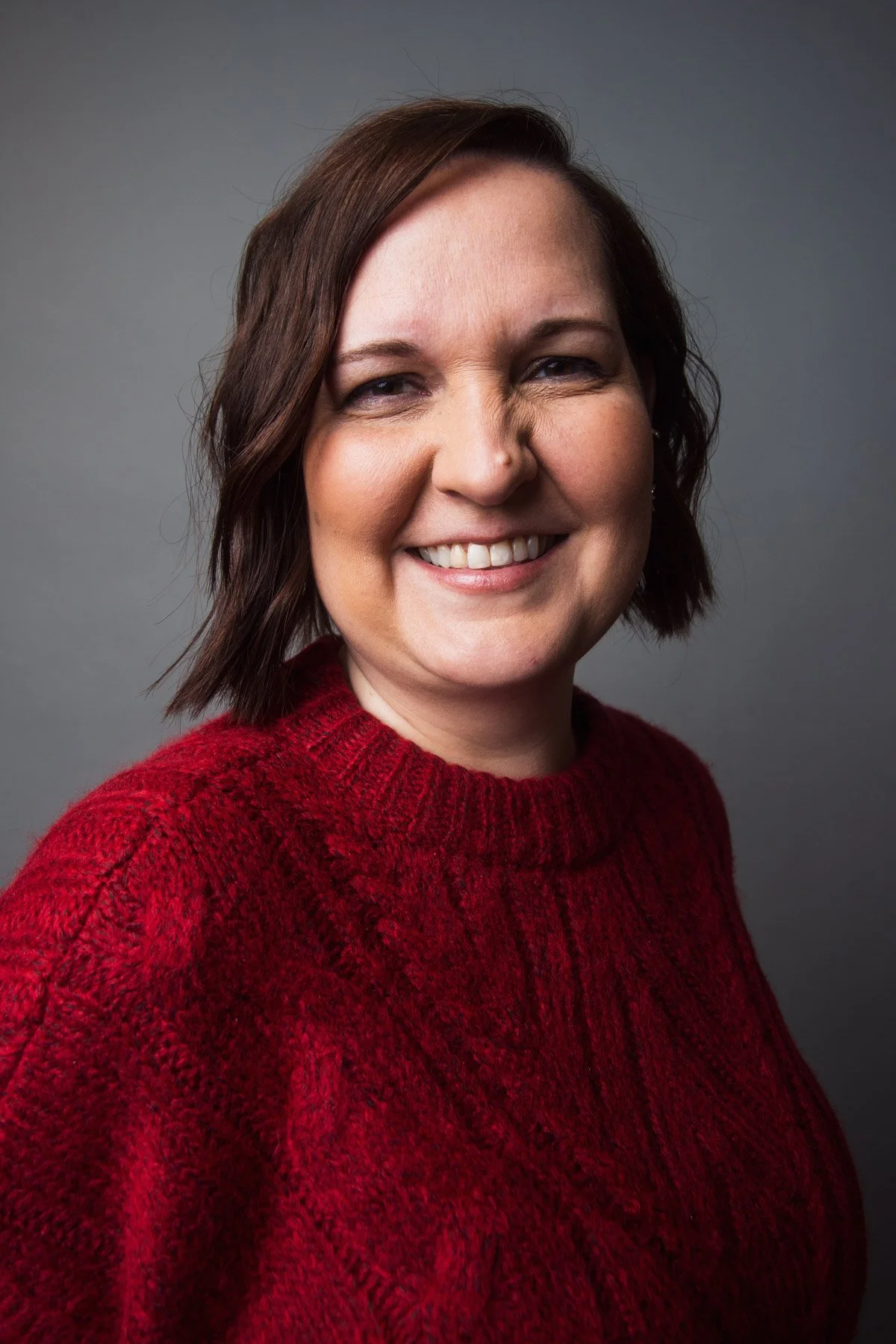 A woman with short brown hair wearing a red knit sweater, smiling against a gray background.