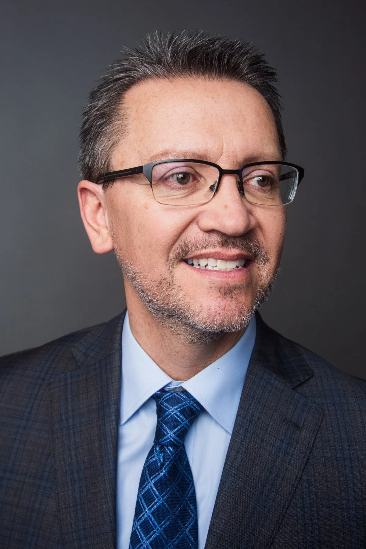 A middle-aged man with glasses, short dark hair, and a beard, wearing a plaid suit and a blue tie, smiling slightly against a dark gray background.