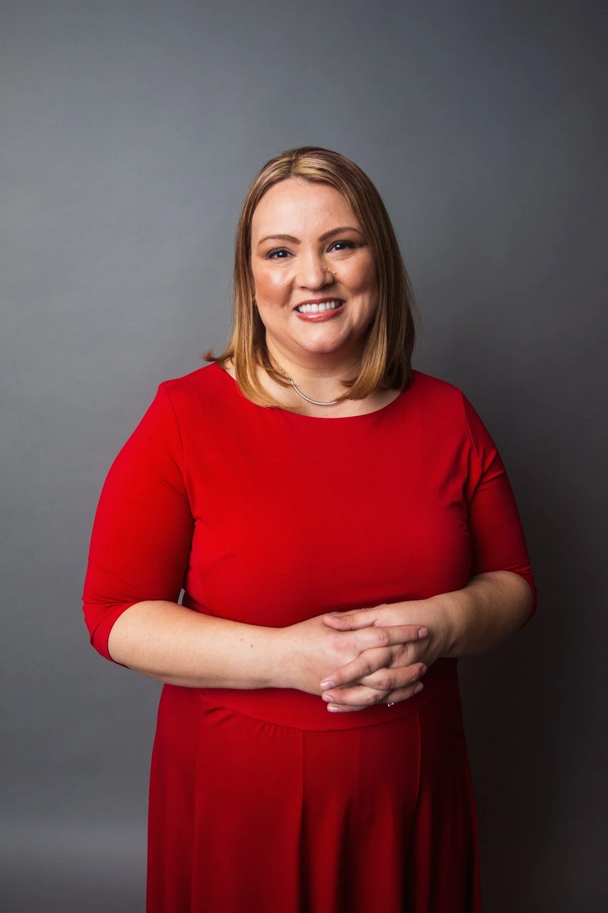 Portrait of a woman with light skin, shoulder-length light brown hair, wearing a red dress, smiling, standing against a gray background.