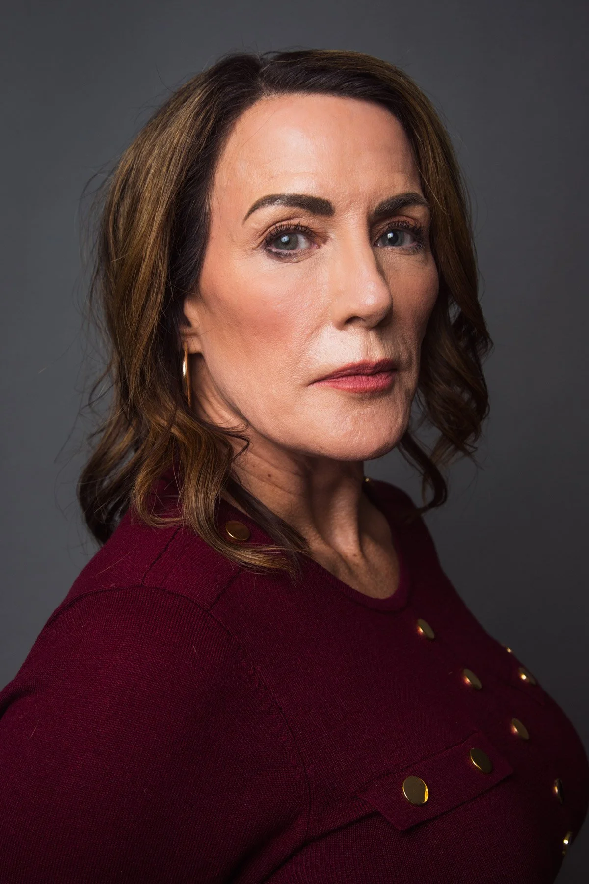 A woman with shoulder-length wavy brown hair, wearing a maroon top with gold buttons, looks directly at the camera with a serious expression, against a neutral gray background.