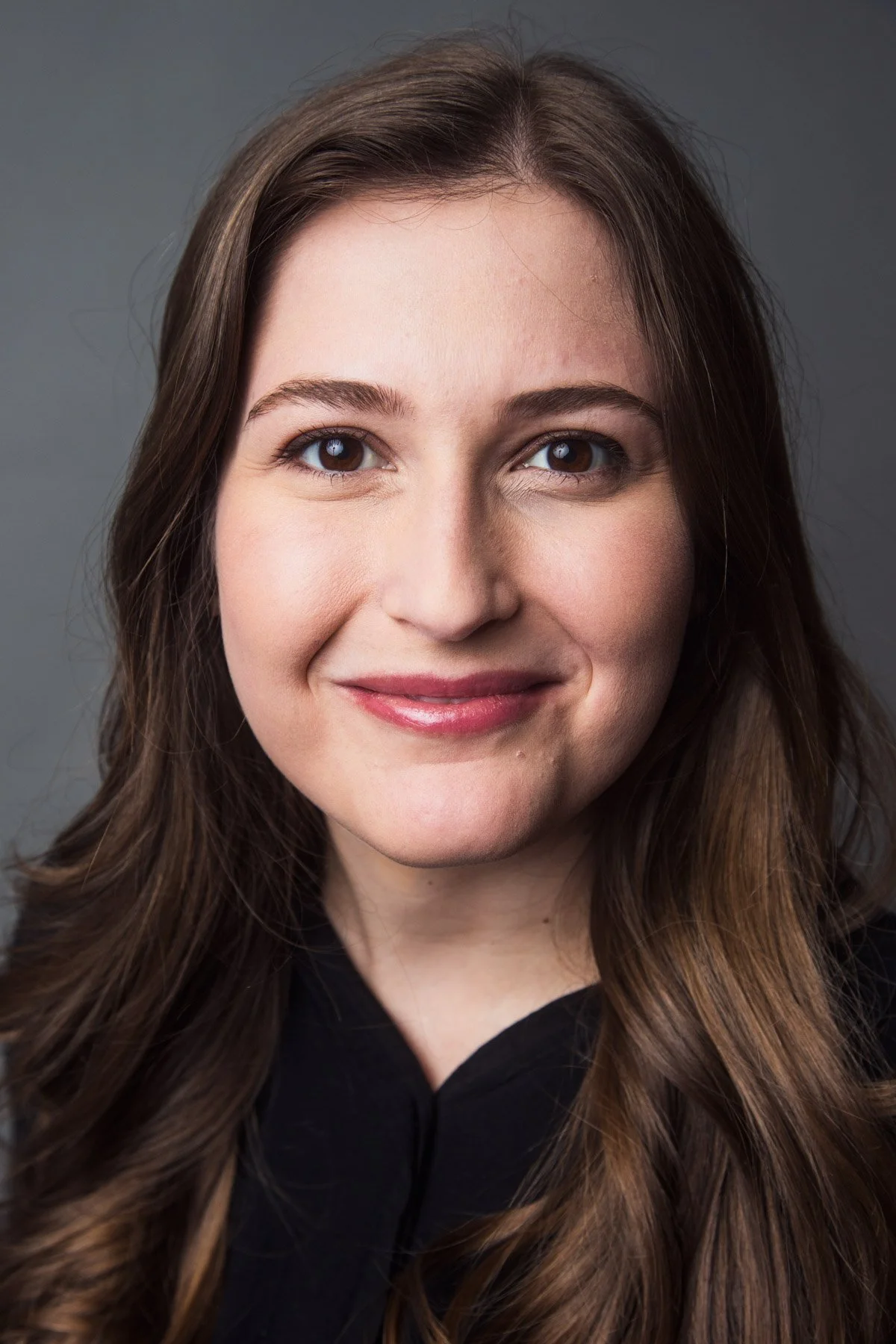 Close-up portrait of a smiling woman with long wavy brown hair and blue eyes, wearing a black top, against a gray background.