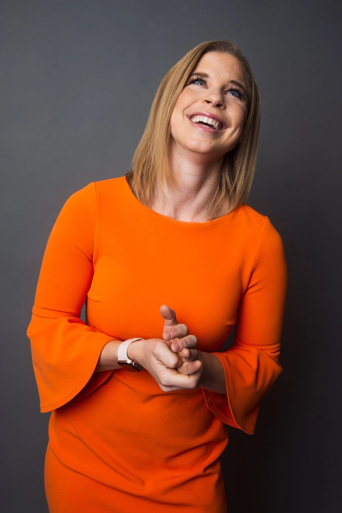 A smiling woman wearing an orange dress with bell sleeves, standing against a dark grey background, with her hands clasped together.