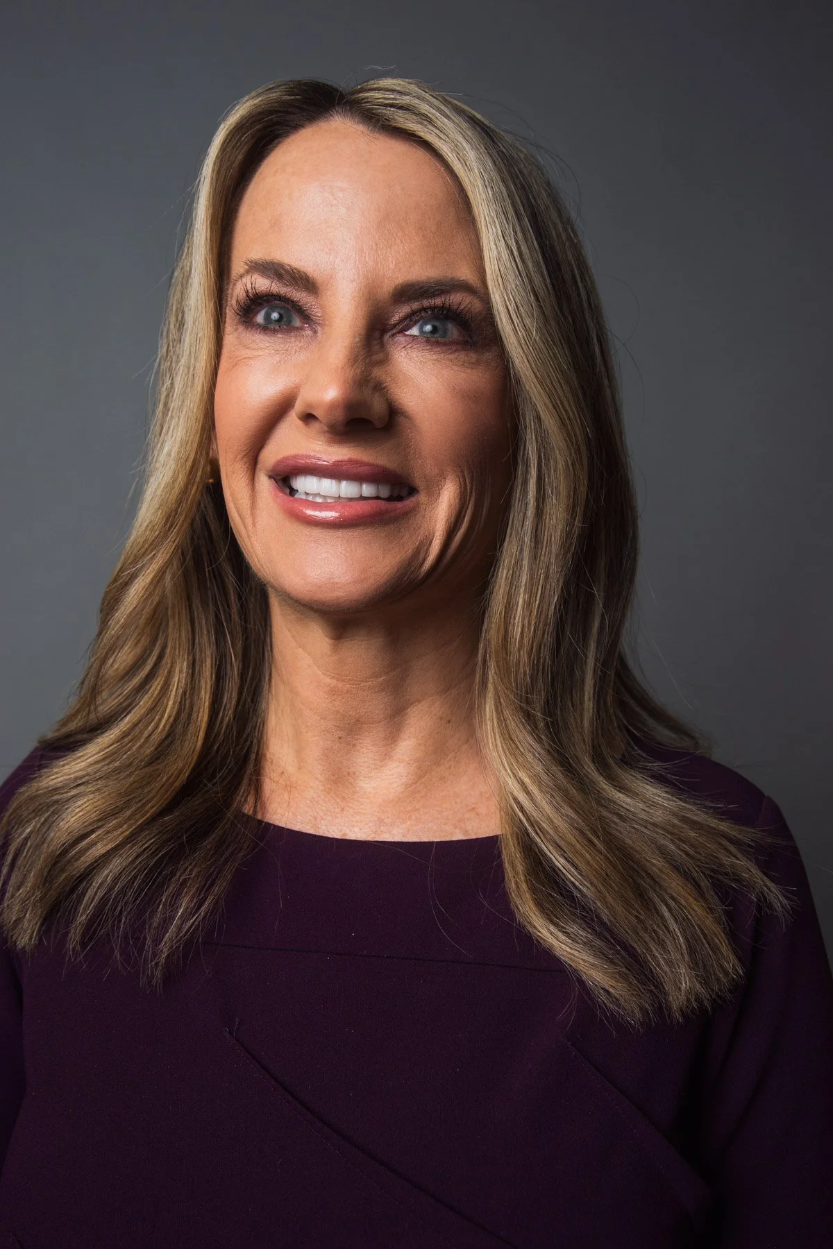 A woman with long blonde hair, blue eyes, and a friendly smile, wearing a dark purple top, against a grey background.