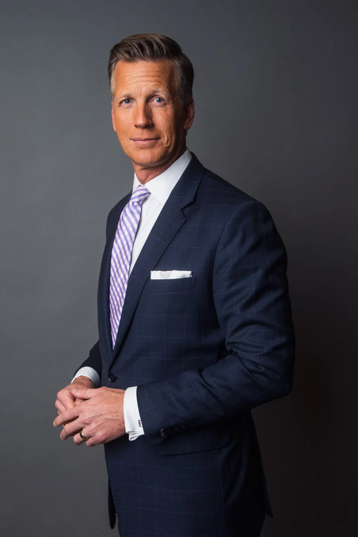 A middle-aged man with light brown hair, dressed in a dark blue checkered suit, white shirt, and purple striped tie, standing against a plain grey background.