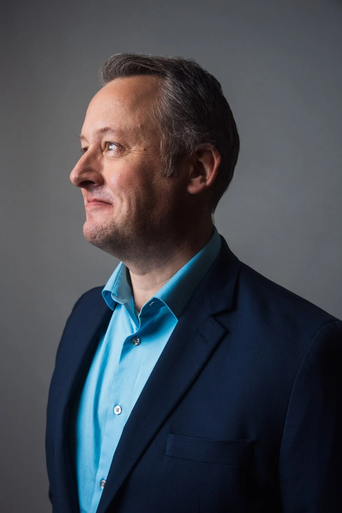 Side profile of a middle-aged man with gray hair in a navy blazer and light blue shirt, smiling slightly, against a gray background.