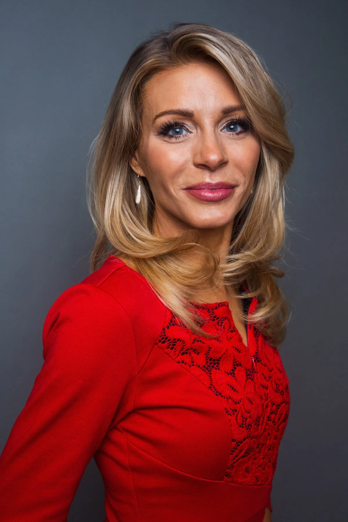 A woman with blonde hair, blue eyes, and light makeup, wearing a red dress with lace detail at the top, standing against a gray background.