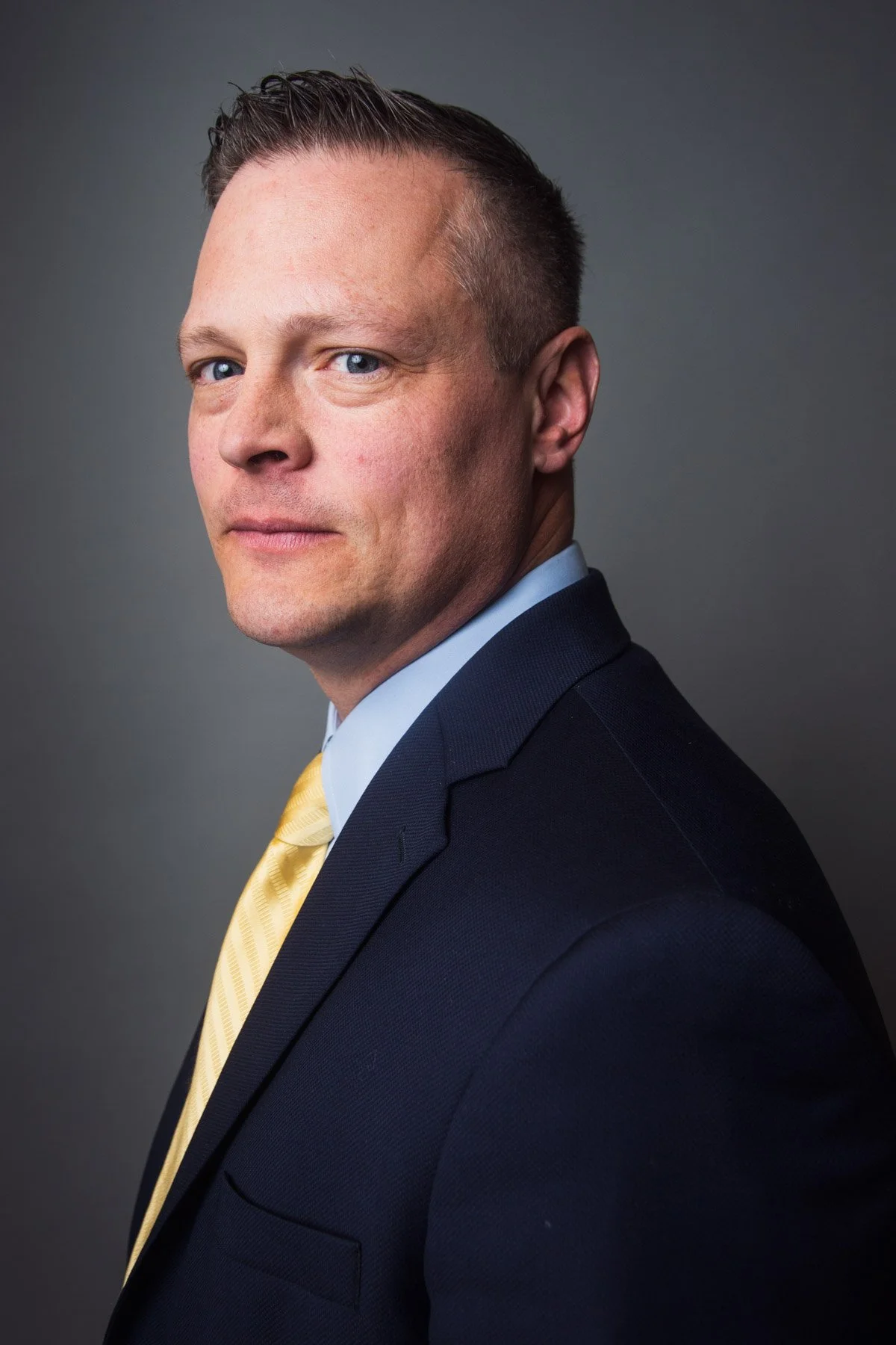 Professional portrait of a man in a dark suit with a light blue shirt and a yellow tie, against a gray background.
