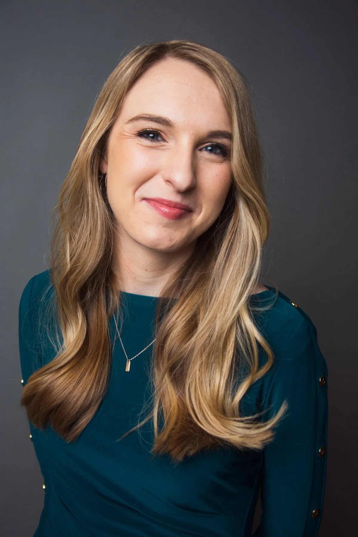 A woman with long, wavy blonde hair, wearing a teal top with gold button details on the sleeves and a silver necklace, smiling against a gray background.