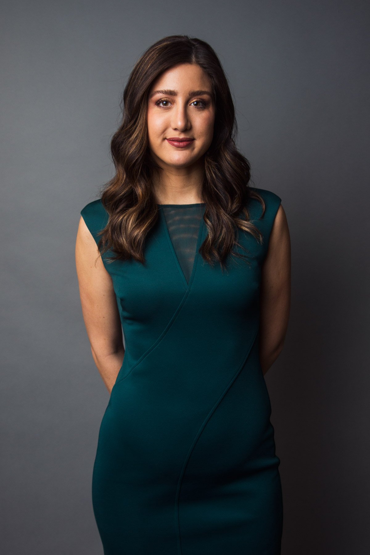 A woman with long, wavy brown hair wearing a teal dress standing against a gray background.