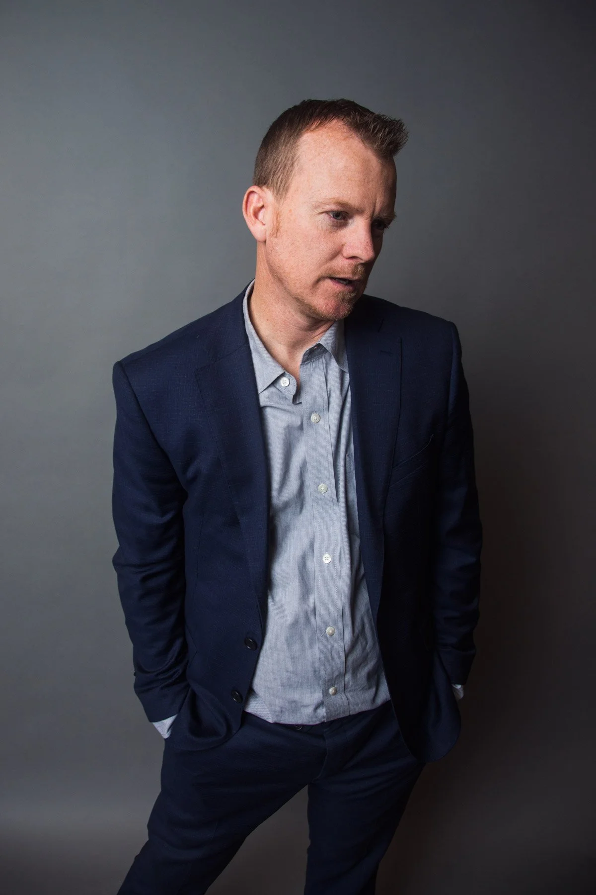 A man with red hair, wearing a dark blue blazer and light blue dress shirt, standing with hands in his pockets, looking down, against a plain gray background.