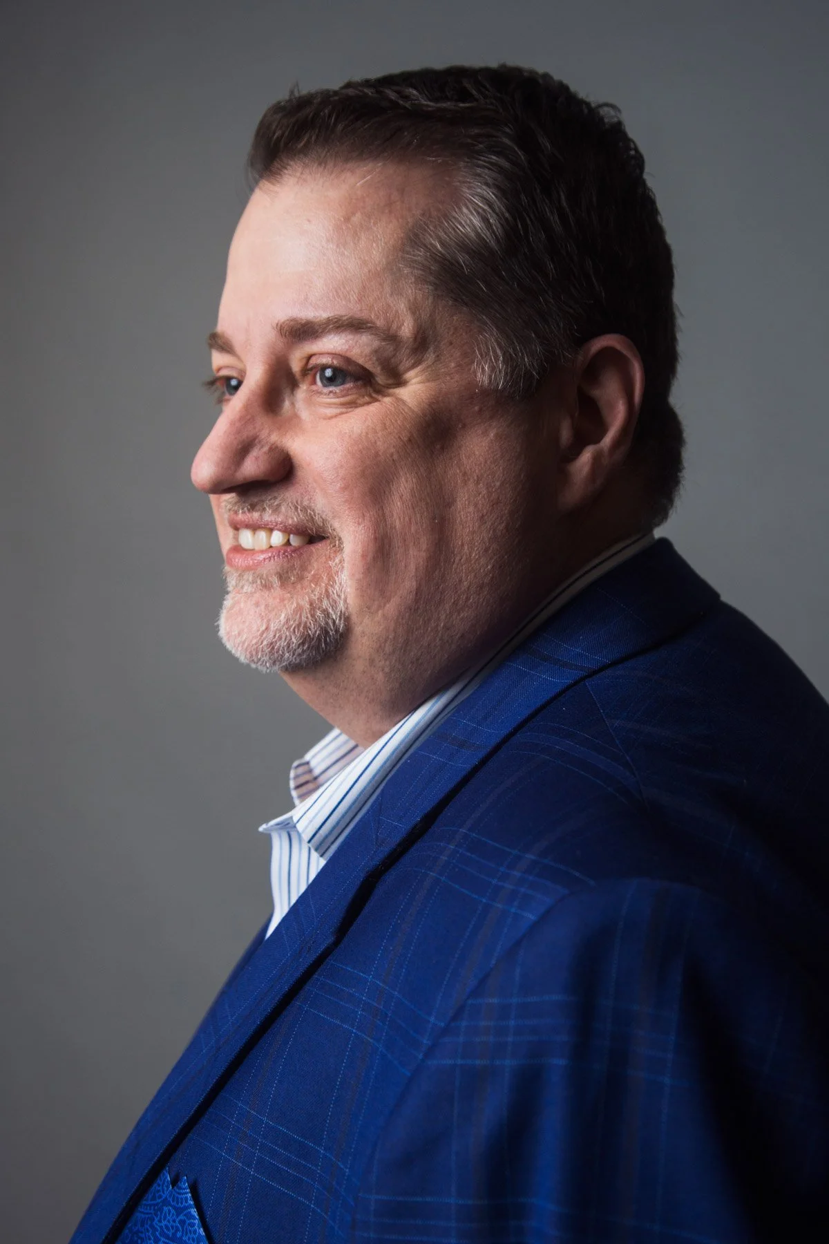 A middle-aged man in a blue plaid suit jacket with a light blue and white striped shirt, smiling, with short dark hair and a goatee with some gray, against a plain gray background.