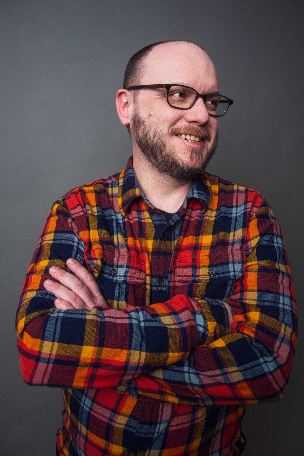 A man with glasses and a beard wearing a colorful plaid shirt, smiling and crossing his arms, against a gray background.