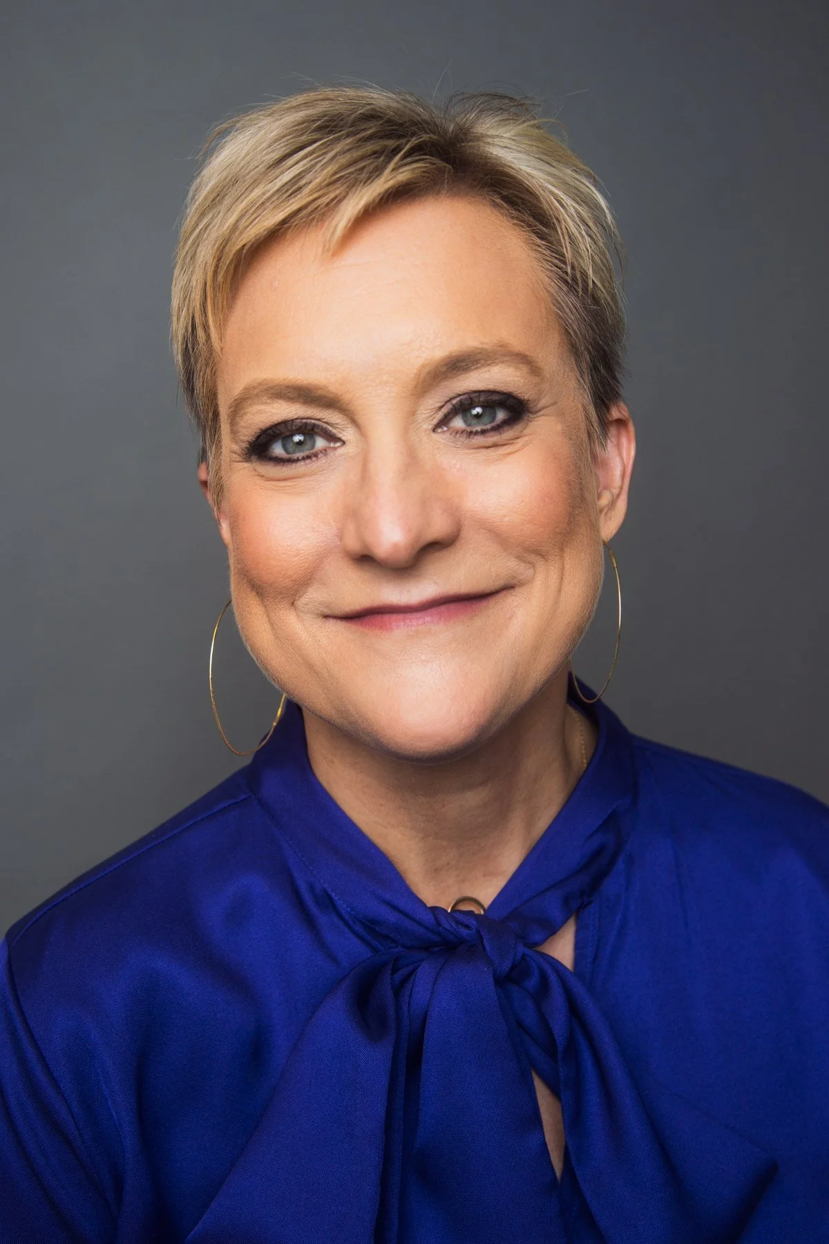 Close-up portrait of a smiling woman with short blonde hair wearing a royal blue blouse with a bow at the neck and large hoop earrings against a gray background.