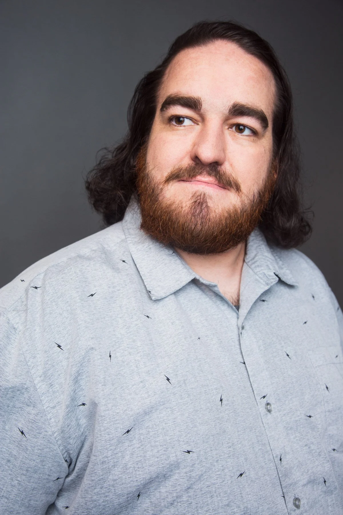 Portrait of a man with long dark hair, beard, and mustache wearing a light gray shirt with small black lightning bolt patterns, against a dark gray background.