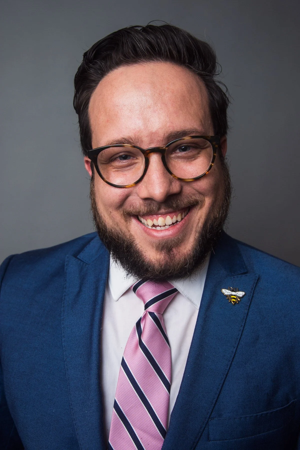Portrait of a man with glasses, a beard, wearing a blue suit, pink and navy striped tie, and a bee lapel pin, smiling against a gray background.