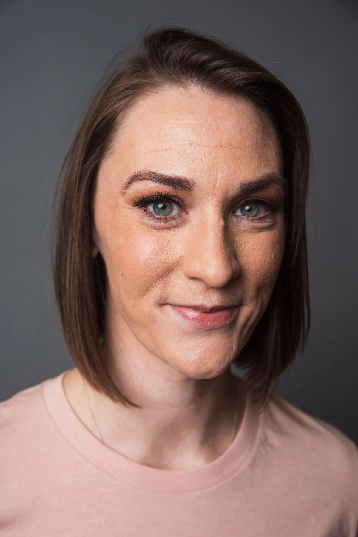 Close-up of a woman with fair skin, shoulder-length brown hair, blue eyes, and a subtle smile, wearing a light pink shirt, against a gray background.