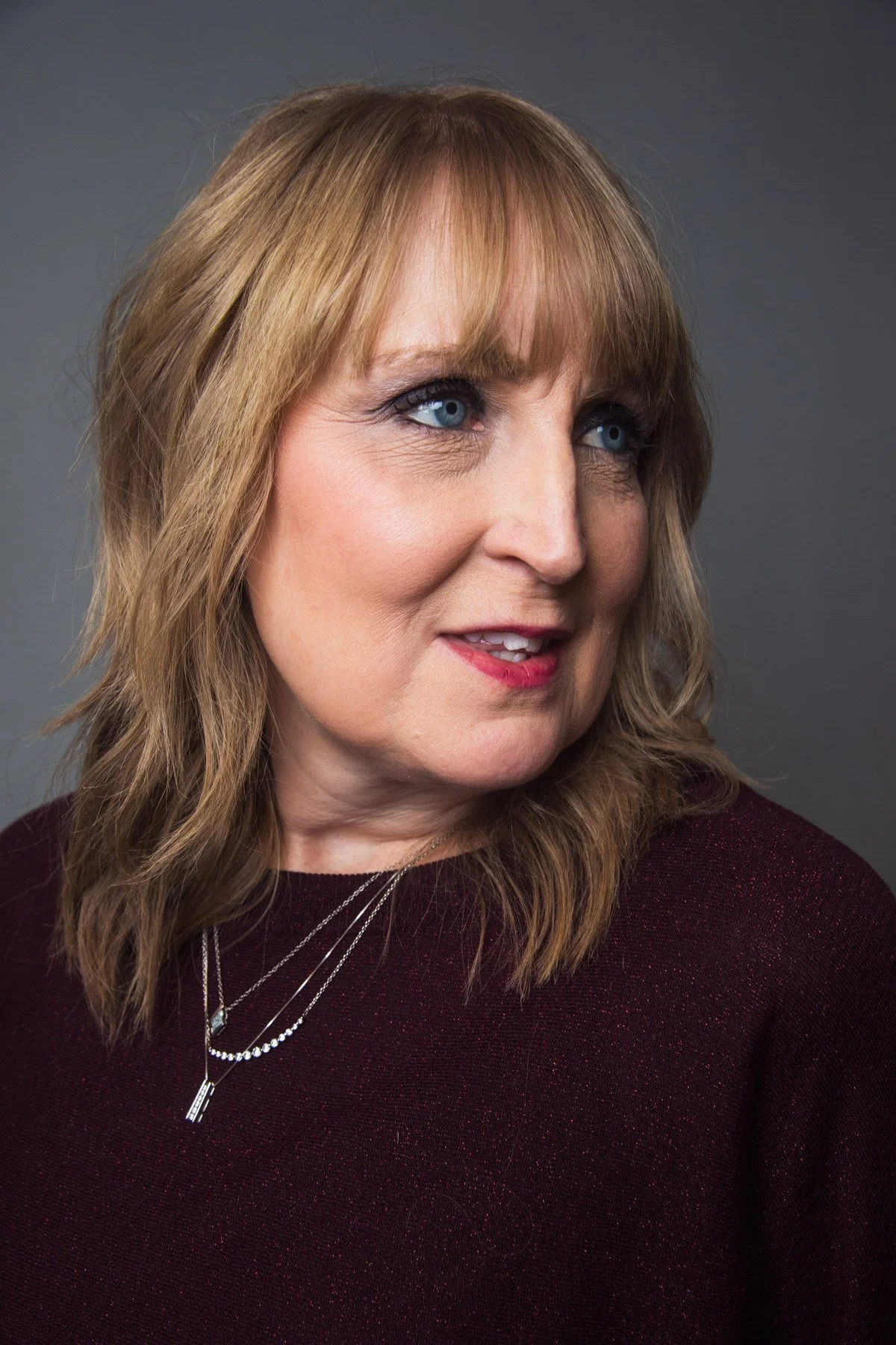 A close-up portrait of a middle-aged woman with shoulder-length blonde hair wearing a dark red top and layered silver necklaces, looking to her right against a neutral gray background.
