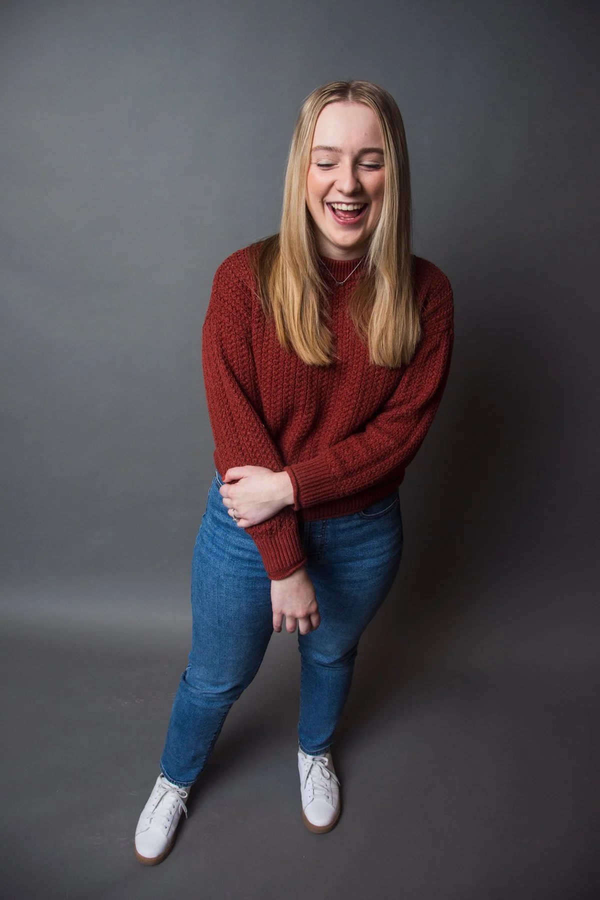 A young woman with long blonde hair laughing, wearing a red sweater, blue jeans, and white sneakers, standing against a gray background.
