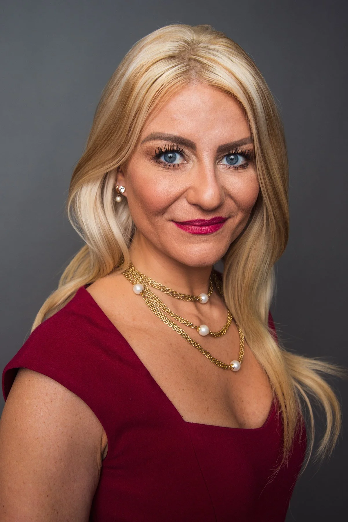 A woman with blonde hair, blue eyes, wearing a red top, gold and pearl necklace, and earrings, smiling at the camera against a gray background.