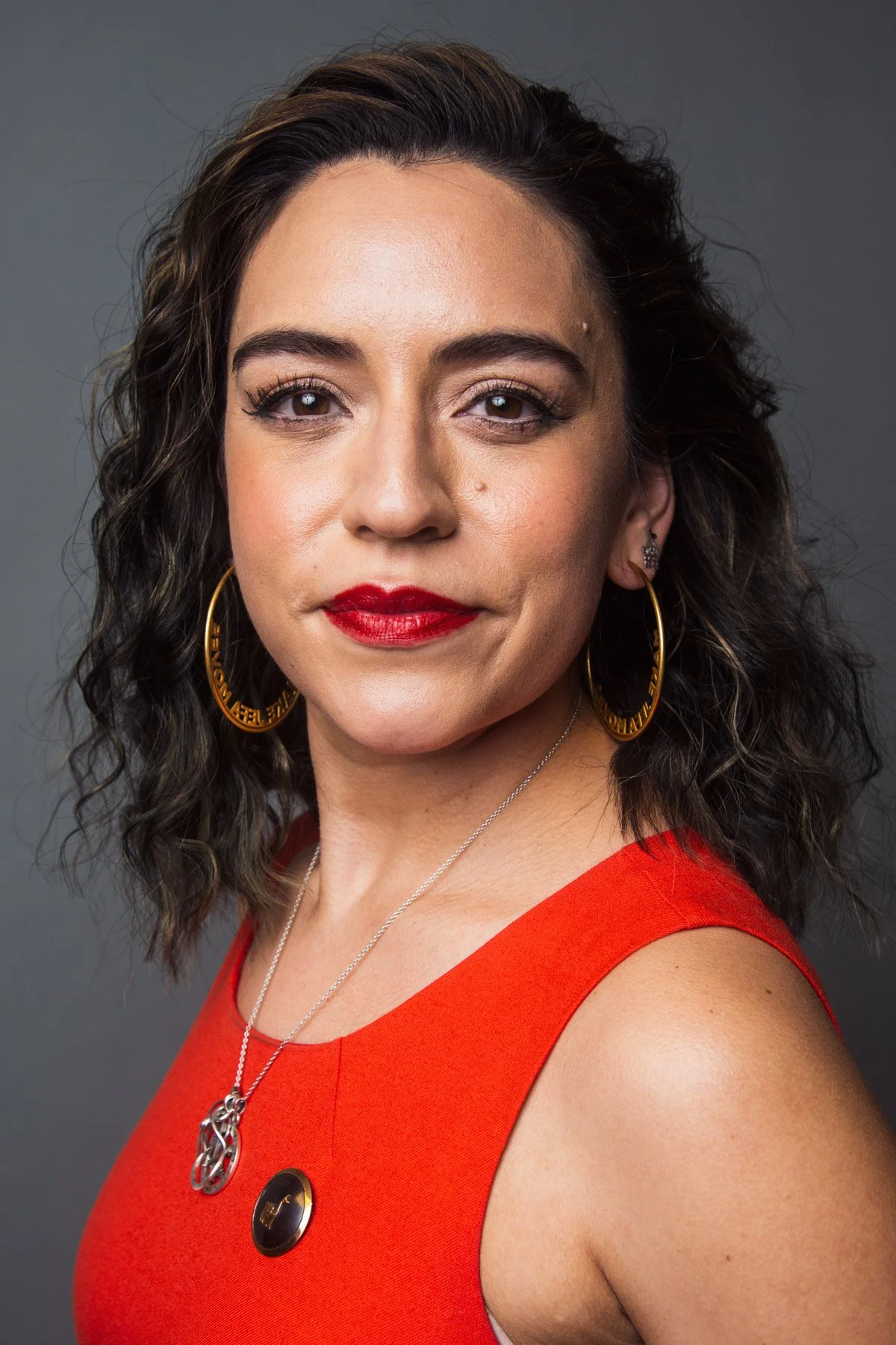 A woman with dark curly hair, wearing red lipstick and hoop earrings, dressed in a sleeveless red top, against a dark grey background.
