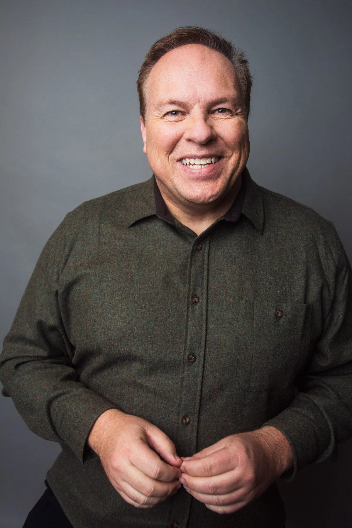 A smiling man with light skin, light brown hair, wearing a dark button-up shirt, standing in front of a plain gray background.