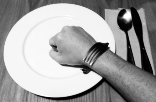 A Liberation Band (i.e. a bent fork) adorns a wrist, while a clenched fist sits atop a plate. Next to the plate is a napkin with a spoon and knife.