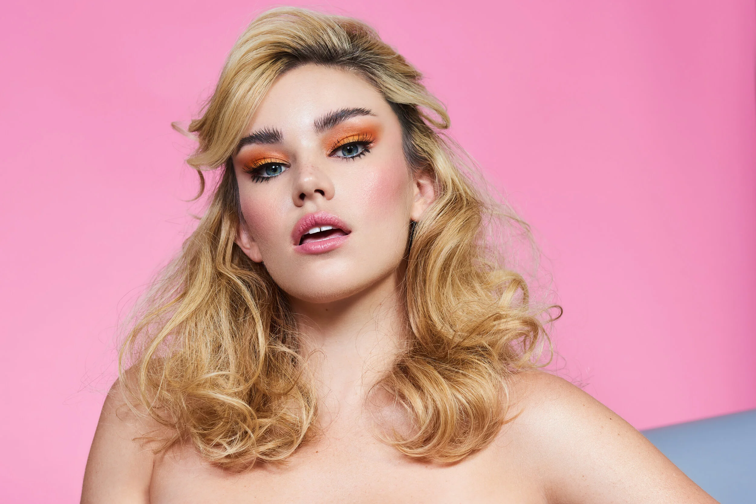 female model in front of bright pink backdrop with big curly hair and orange eyeshadow