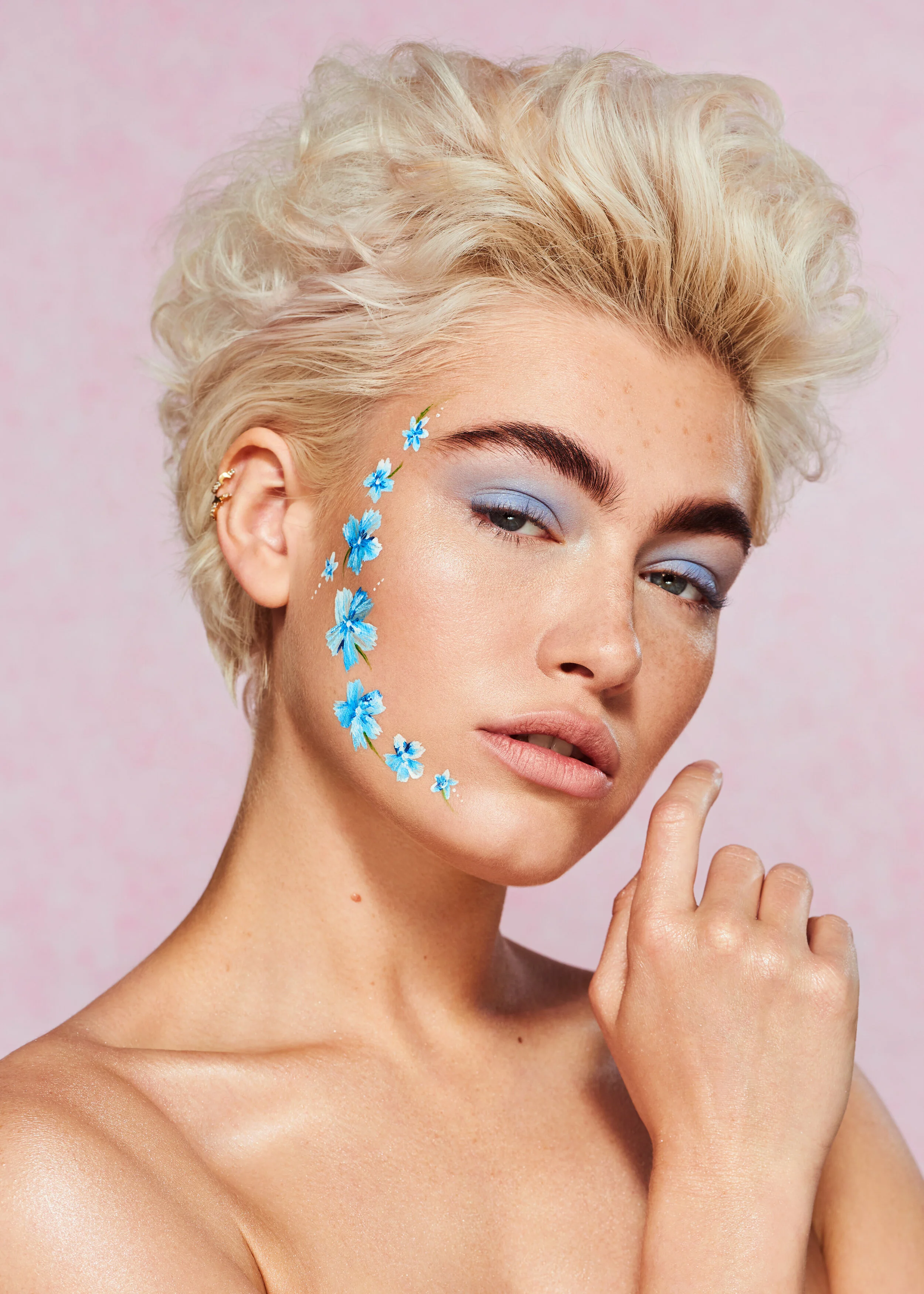 Short hair blonde model with flower face paint art in front of a pink backdrop