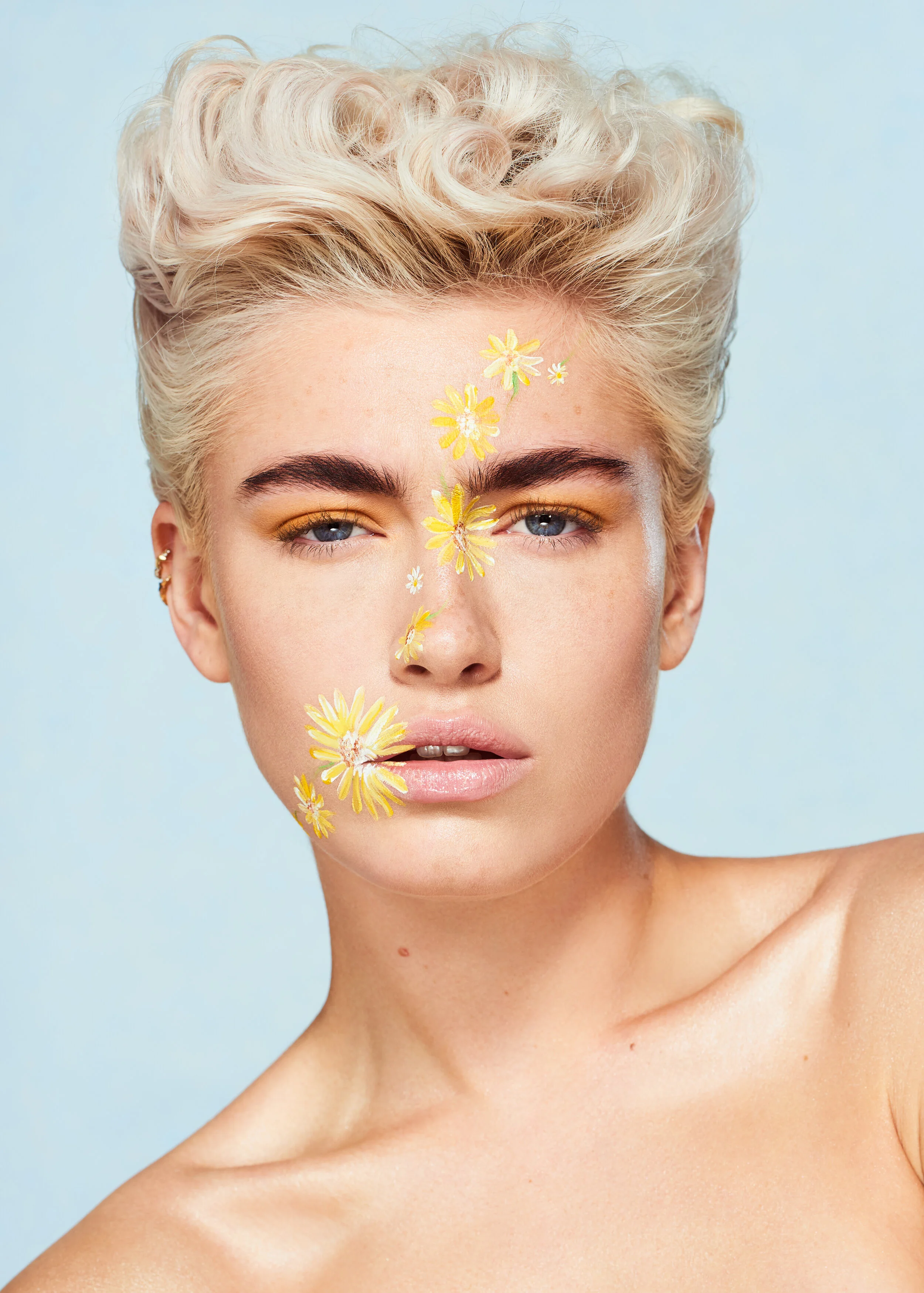 female model with short curly blonde hair and flower face paint art in front of a blue backdrop