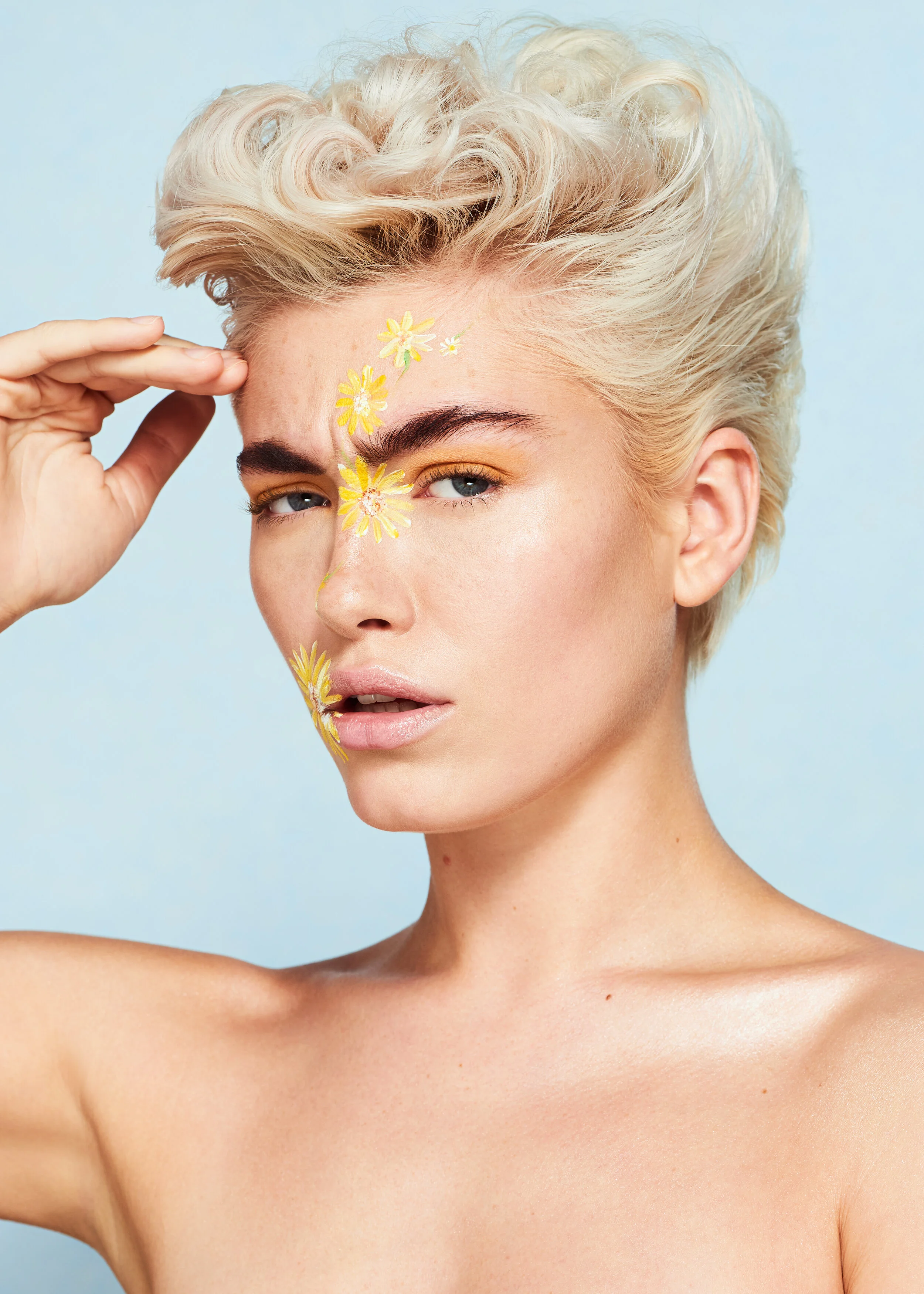female model with short curly blonde hair and yellow flower face paint art in front of a blue backdrop