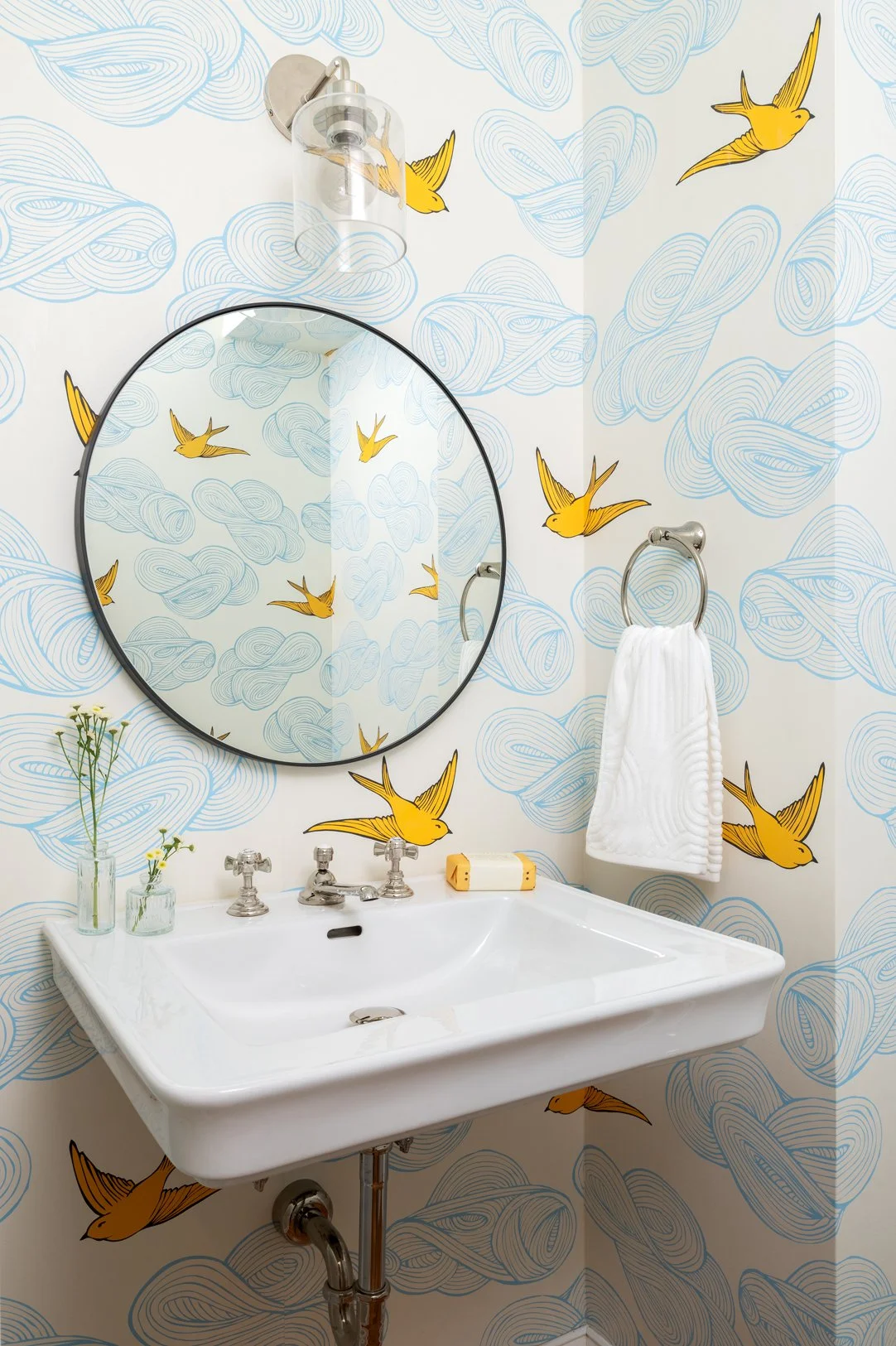 Powder room with white wall-mounted sink and wallpaper with blue clouds and yellow birds.
