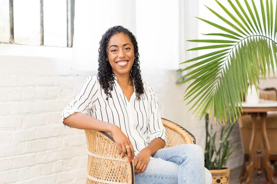 Shila Griffith in a white button-down with black vertical stripes and light blue jeans. She is sitting in a high-bak wicker chair.