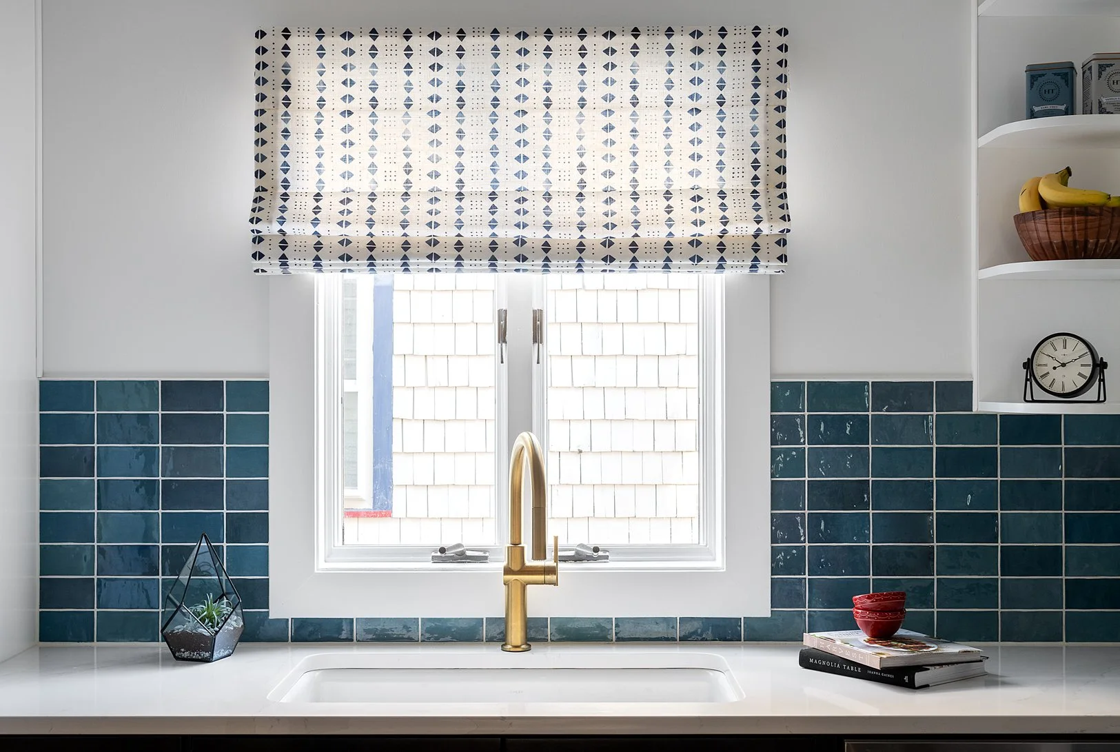 Kitchen window with a patterned roman shade and surrounding blue backsplash.