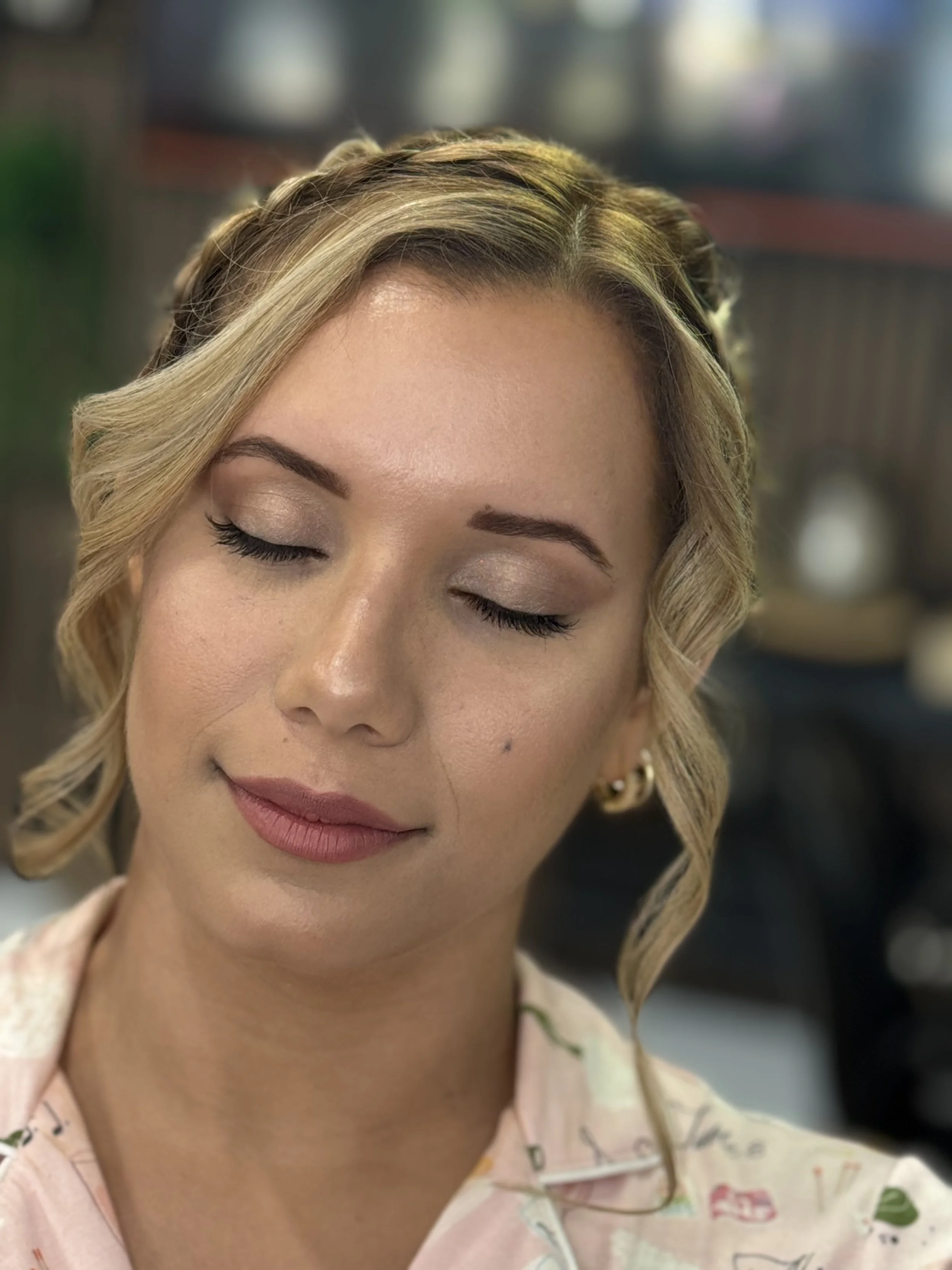 A woman with blond hair styled in soft waves and makeup, with her eyes closed and a peaceful expression, over a blurred indoor background.