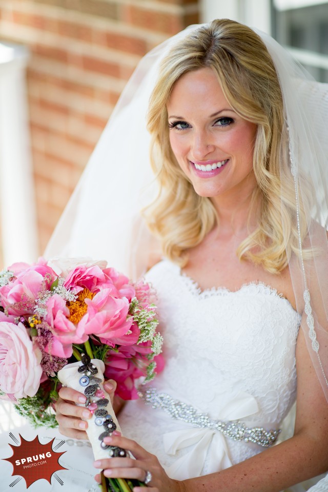 A smiling blonde bride holding a bouquet of pink flowers, wearing a white wedding dress and veil, standing in front of a brick wall.