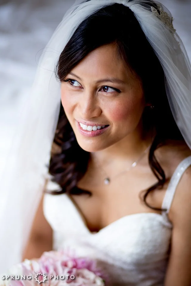 Close-up of a smiling bride with dark hair and a white wedding dress, wearing a veil and holding a bouquet.