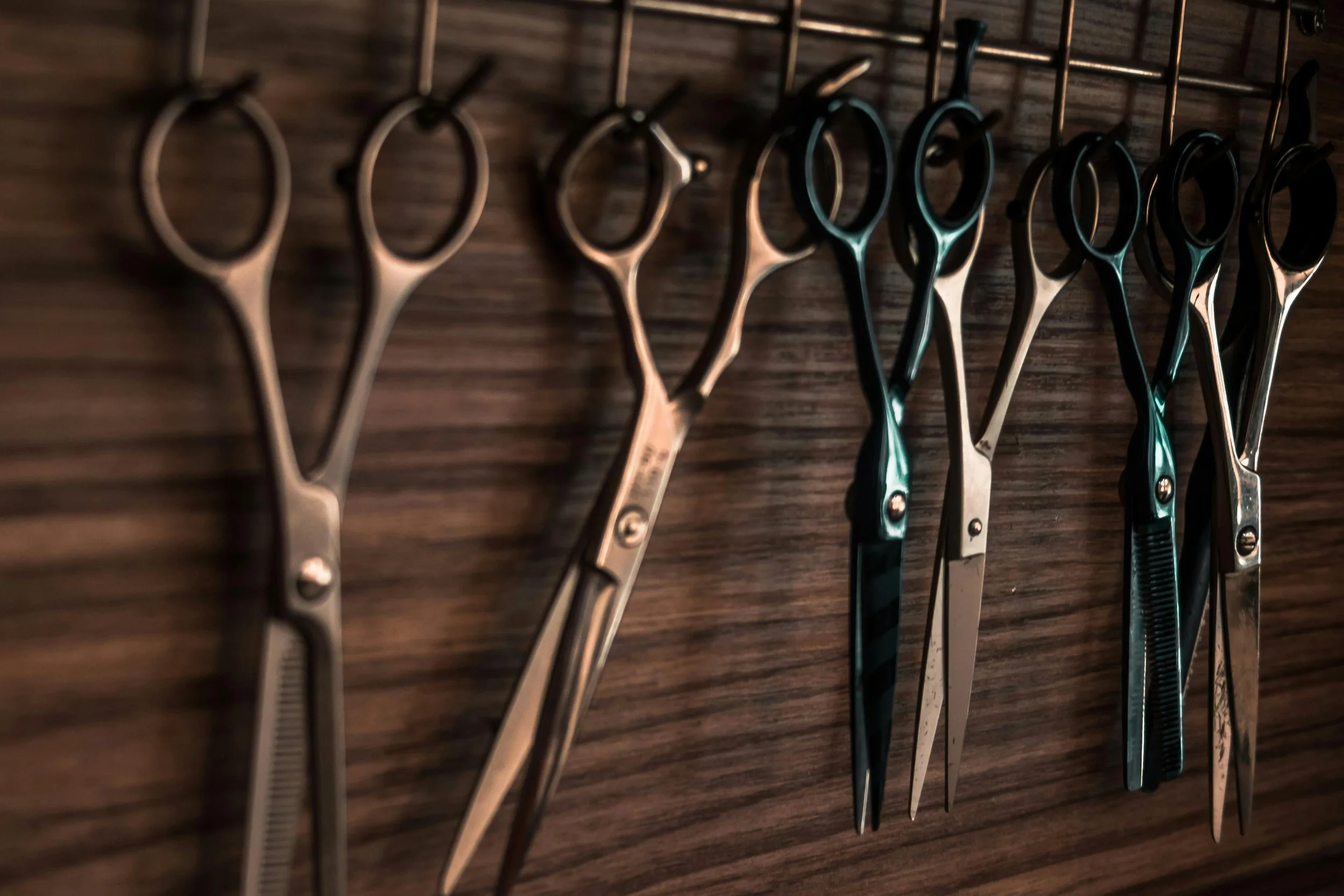 A row of hair scissors hanging on a horizontal metal rod against a wooden wall.