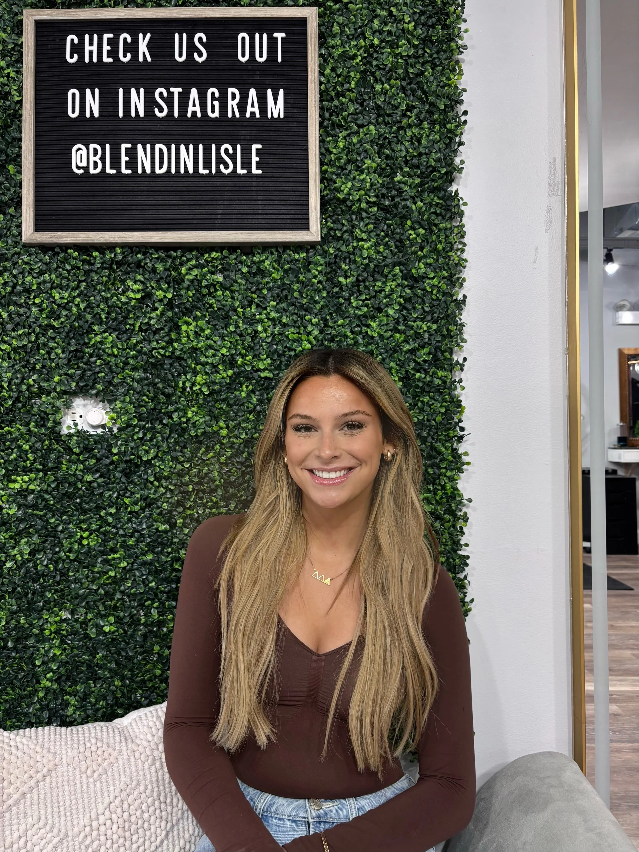 A young woman with long blonde hair smiling, sitting on a beige cushion in front of a green leafy wall. There is a black sign above her with white text that reads "Check us out on Instagram @blendenlisle."