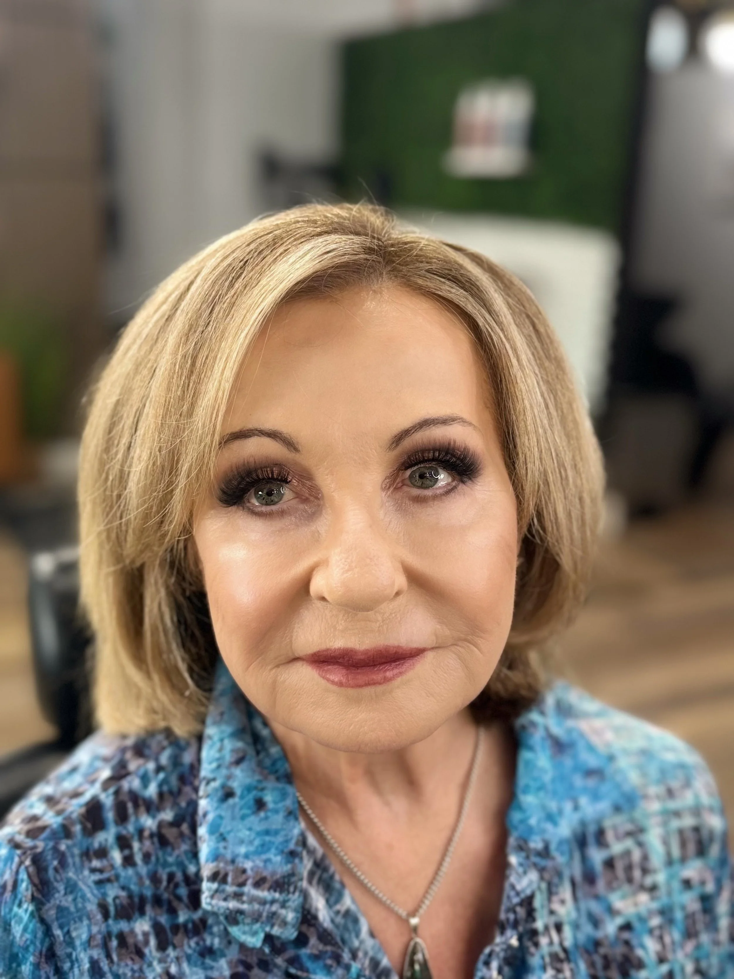 Close-up of a middle-aged woman with blonde hair and makeup, wearing a patterned blue shirt and silver necklace, in a salon or indoor setting.