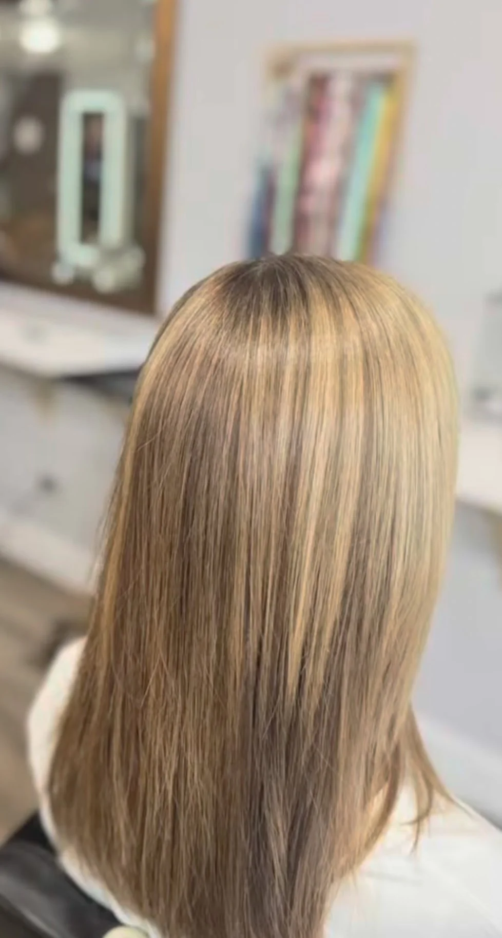 A person with straight, blonde highlighted hair sitting in a salon chair, facing away from the camera, with a mirror and magazines in the background.