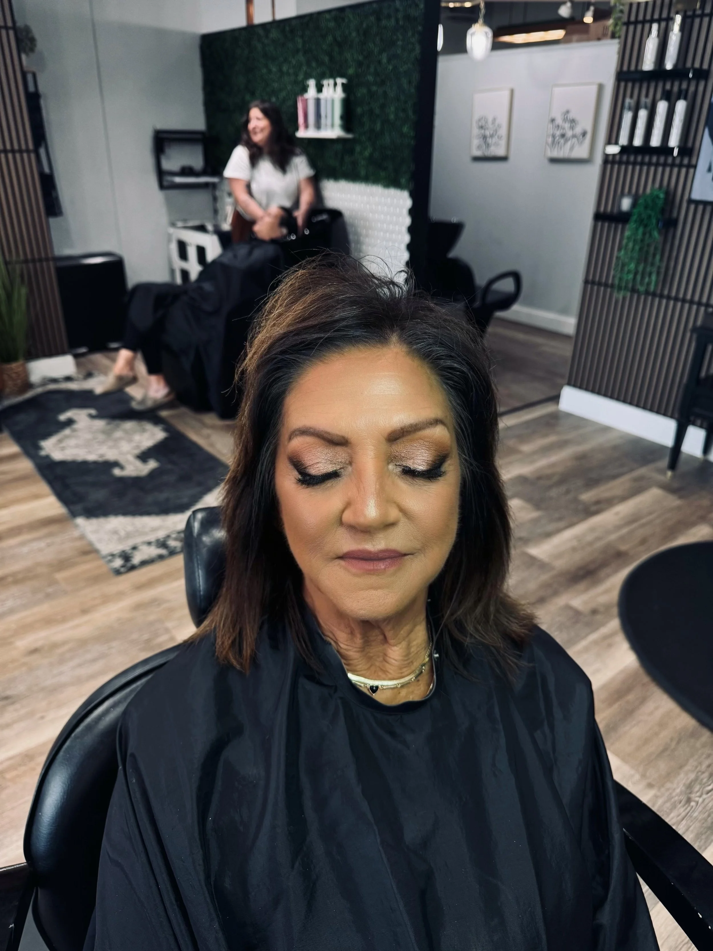 A woman with makeup and dark hair sitting with her eyes closed in a salon chair, wearing a black cape, with a woman in the background smiling and doing another person's hair or makeup in a modern salon.