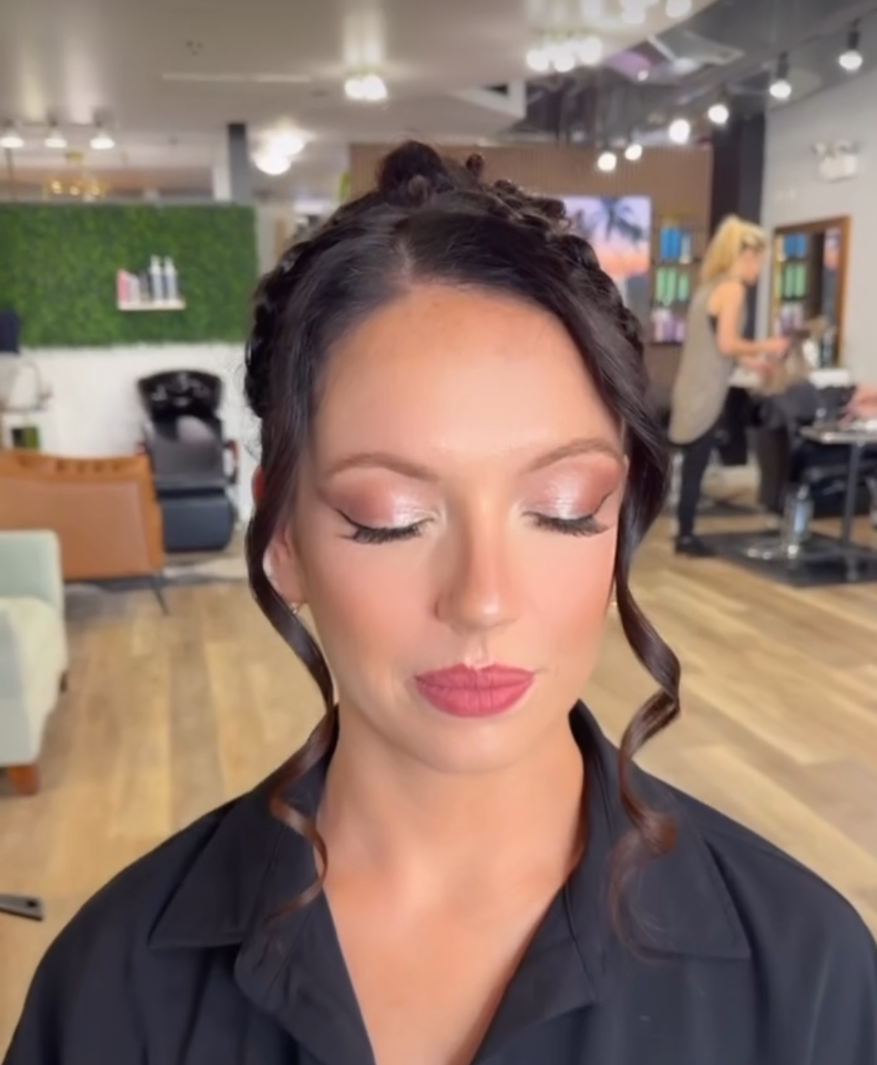 A woman with styled hair and makeup sitting in a salon or beauty studio with hairdresser in background.